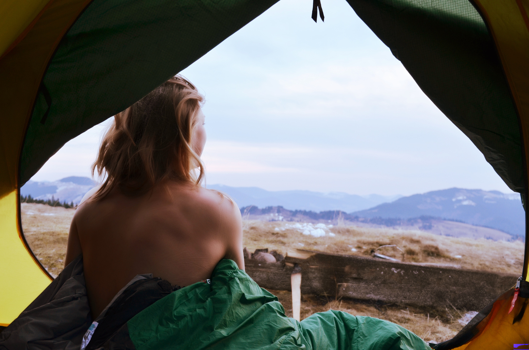 A young woman sitting in the tent with naked back, looking at the mountain landscape in winter
